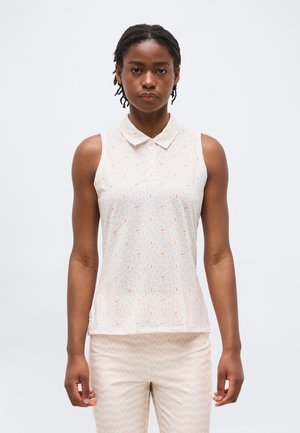 Žena s krátkymi vrkočmi, oblečená v bielom bezrukávovom top s golierom so žltými malými vzormi a svetlo béžových nohaviciach.