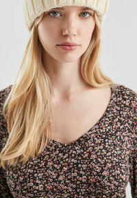 Bloemenpatroon top in donkere tinten met een V-hals. Zachte stof met kleine bloemen in wit, geel en rood. Crème gebreide muts voegt textuur toe.