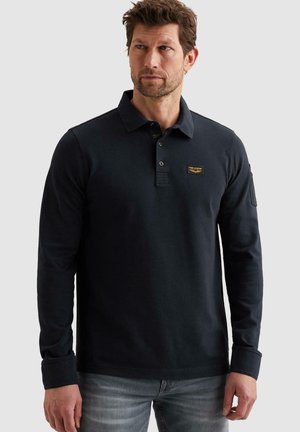 Uomo con capelli corti e barba, indossa una polo nera a maniche lunghe con colletto abbottonato e jeans grigi, in piedi davanti a uno sfondo semplice.