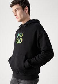 Sudadera negra con un bolsillo frontal y capucha con cordón. Presenta un logo reflectante "HUGO" en tonos verdes y amarillos. Tejido suave, ajuste casual.