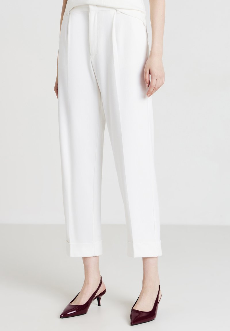 Donna che indossa pantaloni bianchi a vita alta e tagliati con pieghe frontali e décolleté in camoscio borgogna con punta a punta su un pavimento bianco.