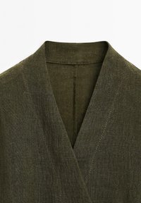 Vêtement vert olive avec un col châle, fabriqué à partir d'un tissu texturé. Présente un design enveloppant et des coutures visibles le long des bords.