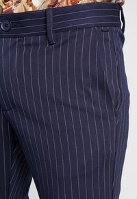 Pantalons en navy à rayures fines en tissu lisse, avec une fermeture à bouton, des poches latérales et des rayures et coutures blanches contrastantes.