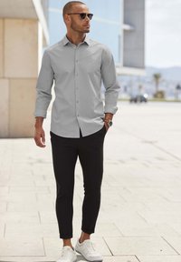 Helles graues Hemd mit Knopfleiste, schwarze schmal geschnittene Hose und weiße Sneakers; das Model trägt Sonnenbrille und steht im Freien auf einem gefliesten Boden.