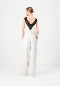Sleeveless cream top features a deep black lace back detail; paired with loose, high-waisted, textured white pants.