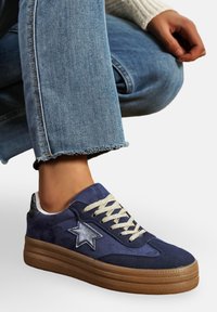Personne portant des sneakers bleu en daim avec des étoiles, des lacets beiges et des semelles en plateforme marron, assorties à un jean bleu avec un ourlet effiloché.