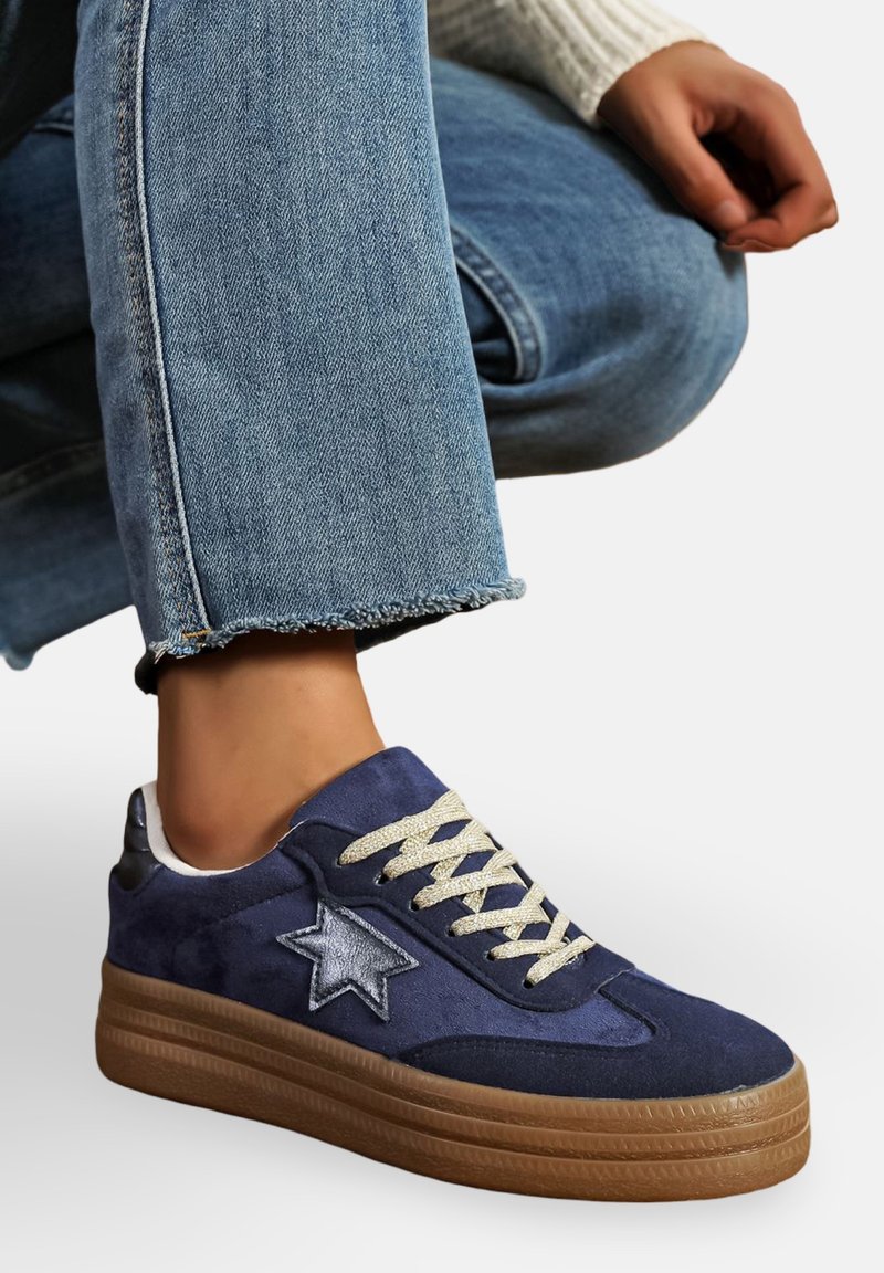 Personne portant des sneakers bleu en daim avec des étoiles, des lacets beiges et des semelles en plateforme marron, assorties à un jean bleu avec un ourlet effiloché.