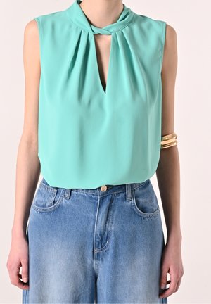 Ærmeløs tealbluse med rynket halsudskæring og lille frontudskæring, stoppet ned i højtaljede lyseblå cowboybukser, gyldne armbånd på armen.