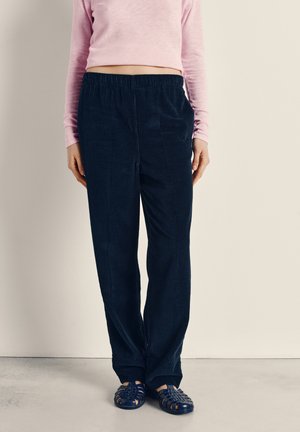 Osoba ubrana w granatowe sztruksowe spodnie z elastycznym pasem, jasnoróżową bluzkę z długim rękawem oraz granatowe plecione sandały, stojąca na tle jednolitej ściany.