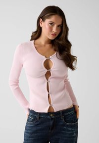 Růžový kardigan s žebrovaným vzorem a dlouhými rukávy, s průstřihem na klíč a ozdobnými knoflíky. Nošený přes tmavě modré džíny. Hladká textura.
