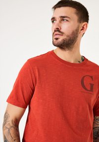 Camiseta roja de manga corta hecha de algodón suave; cuenta con un cuello redondo y un logo negro "G" en el pecho. Ajuste casual y relajado.