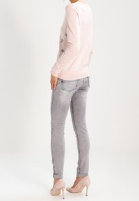 Femme portant un pull rose clair imprimé d'étoiles, un jean skinny gris et des escarpins roses assortis, debout devant un fond clair uni.