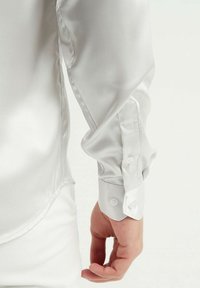 Chemise blanche en satin avec une texture lisse, des poignets classiques à boutons et un ourlet arrondi. Gros plan sur le poignet et la manche.