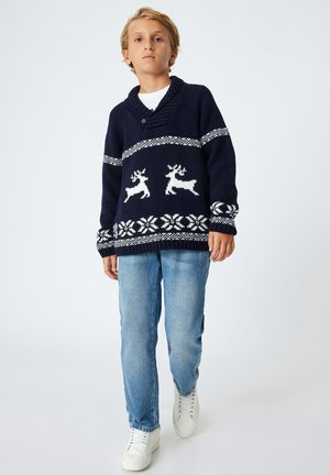 Maglione lavorato a maglia navy con motivi di renne bianche e fiocchi di neve, con scollo a V e polsini a costine, abbinato a jeans azzurri chiari e scarpe bianche.