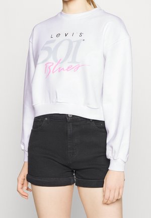 Osoba ubrana w białą, długą bluzę typu crop z napisem „Levi's 501 Blues” oraz czarne, podwinięte szorty dżinsowe.