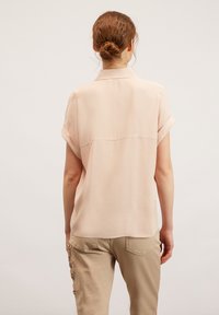 Světle růžová blůza s krátkým rukávem a límečkem, hladká textura, detaily švu na zadním díle, spárována s béžovými kalhotami se zdobnými prvky.