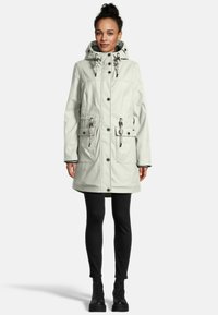 Vrouw in een lichtgrijze capuchonjas met trekkoorden, zwarte leggings en zwarte veterschoenen, staand tegen een eentonige achtergrond.