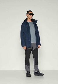 Veste imperméable bleu marine avec capuche, chemise grise et pantalon bicolore noir et gris. Le mannequin porte des lunettes de soleil et des chaussures de sport noires.