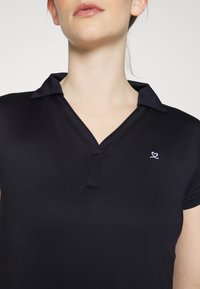 Svart pikétröja i slätt tyg med krage, prydd med ett litet lila broderat hjärtlogga på vänster bröst.