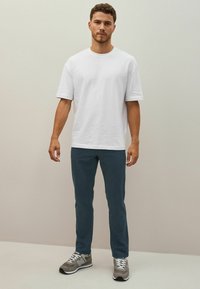 Tricou alb din bumbac, cu o croială relaxată, asortat cu pantaloni de culoare verde închis. Adidași gri cu șireturi albe și o talpă texturată.