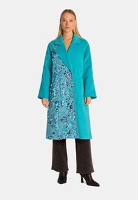 Türkiser Mantel mit einem Schalkragen und blumigem Muster entlang der Vorderseite; getragen über dunklen Hosen mit glitzernder Textur.
