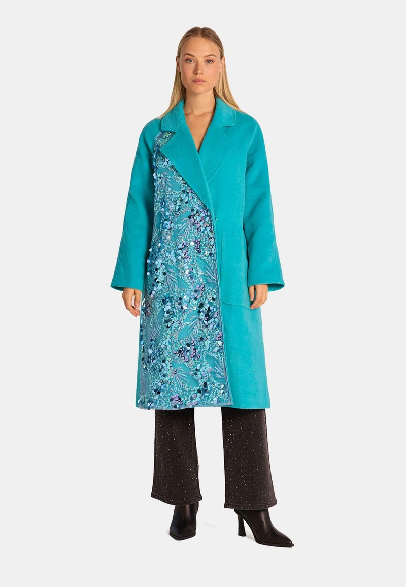 Türkiser Mantel mit einem Schalkragen und blumigem Muster entlang der Vorderseite; getragen über dunklen Hosen mit glitzernder Textur.