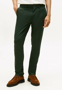 Pantaloni tagliati verde scuro realizzati in tessuto texturizzato, con chiusura a bottoni e tasche laterali, indossati con scarpe in suede marroni.