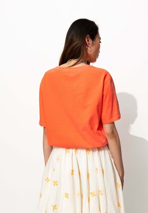 T-shirt orange à manches courtes avec une coupe décontractée, doté d'un large décolleté. Porté sur une jupe blanche à motifs floraux jaunes.