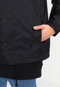 Svart nylonjacka med dragkedja framtill, elastiska muddar och sidoficka. Bärs över en svart sweatshirt med dragsko i nederkanten.