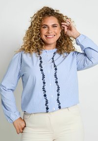 Camisa de botón a rayas azul claro con bordado floral marino en el centro y cuello con volantes; mangas largas recogidas en las muñecas.