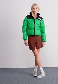 Zelena pufasta jakna s crnim ovratnikom, s prednjim zatvaračem, uparena s smešnom suknjom koja ima prednji prorez, bijelim čarapama i kariranim slip-on cipelama.