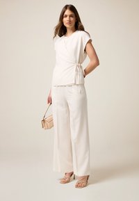 Top bianco avvolgente con maniche corte e cintura laterale, abbinato a pantaloni bianchi a gamba larga. La modella tiene una borsa color beige chiaro ed è vestita con tacchi a strap.
