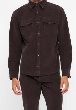Homme portant une chemise en velours côtelé marron foncé boutonnée avec poches poitrine à rabat et pantalon en velours côtelé assorti sur fond uni.