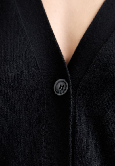 Cardigã preto com um design de decote em V, feito de tecido de malha texturizado, com um único botão preto na abertura frontal.