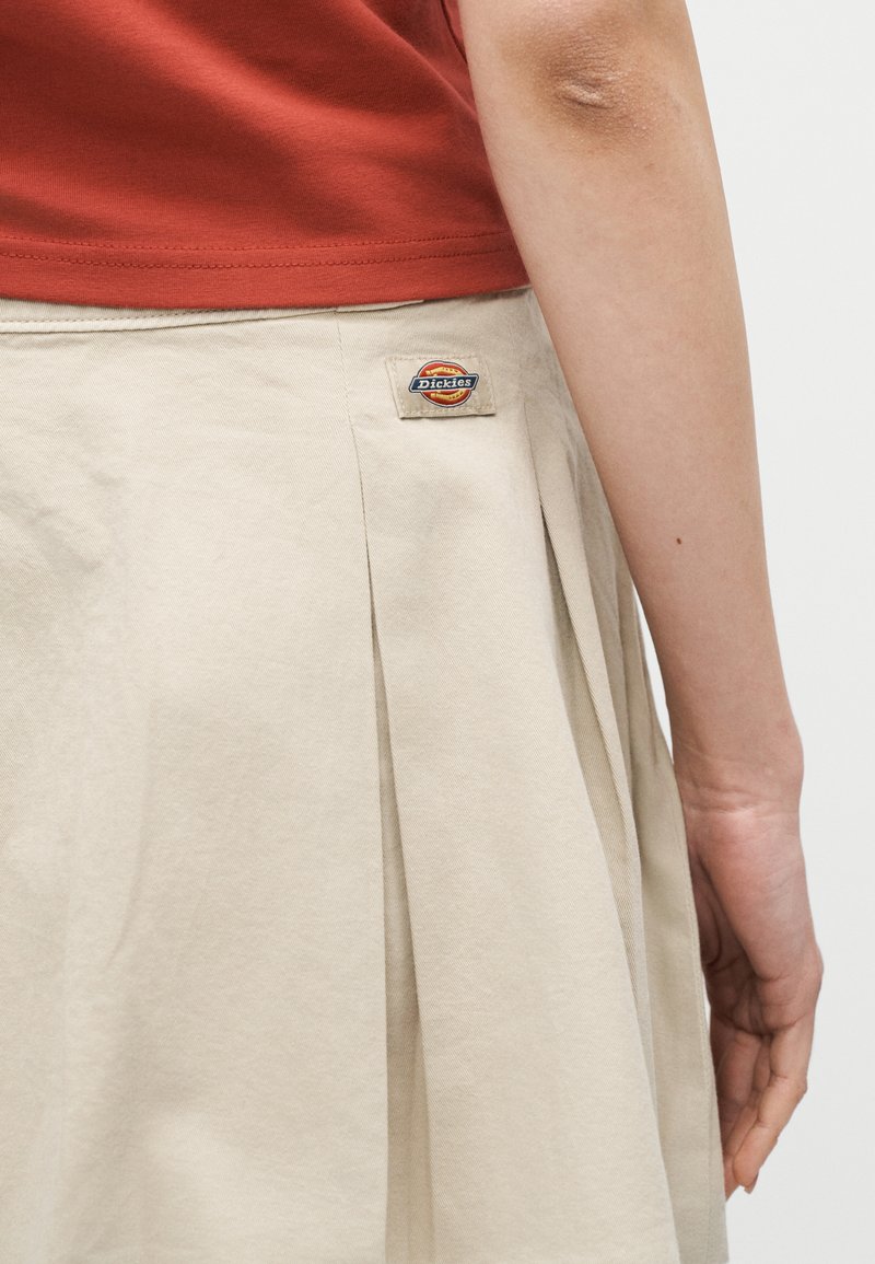 Gonna beige plissettata con logo Dickies indossata con top arancione bruciato, mostrando il braccio destro e la mano contro uno sfondo chiaro.