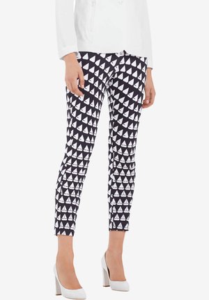 Une personne porte un pantalon noir à motif de voiliers blancs répétés, une veste blanche et des chaussures blanches à talons hauts, debout.