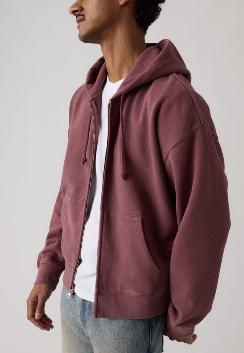 Man draagt een bordeauxrode zip-up hoodie met trekkoorden en zakken aan de voorkant over een wit T-shirt en blauwe spijkerbroek, staand in zijaanzicht.