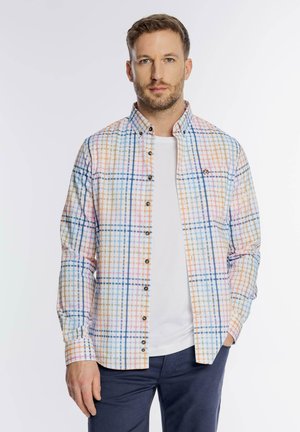 Man wearing multicolored plaid button-up shirt over white t-shirt, standing with one hand in navy pants pocket against plain background.