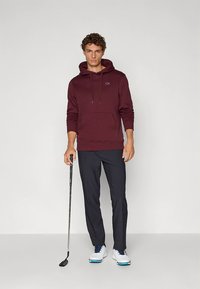 Camisola com capuz bordeaux com bolso frontal e cordões. Combinada com calças escuras e sapatos de golfe brancos. Segurando um taco de golfe. Visual limpo e casual.
