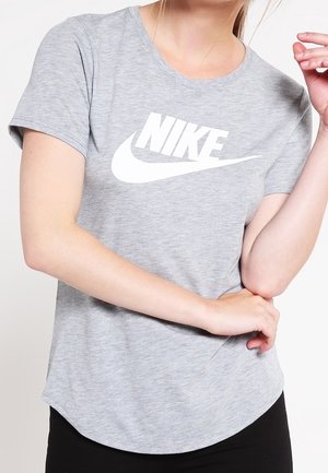 Osoba ubrana w jasnoszarą koszulkę Nike z białym logo, z założonymi na wysokości talii rękami, stojąca na jednolitym tle.