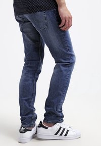 Mörkblå denimjeans för män med slim fit, med subtila slitage och en klassisk femfickdesign, parat med vita och svarta sneakers.