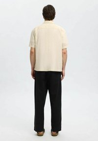 Camicia a maniche corte di un leggero beige con motivo ad occhiello, abbinata a pantaloni neri larghi. Piedi in sandali marroni, visti da dietro.