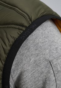 Gepolsterte olivgrüne Weste mit grauem Unterlayer, ausgestattet mit einem schwarzen Gummiband am Hals mit dem Text "Pure Legend". Texturierte Gewebedetails sind sichtbar.