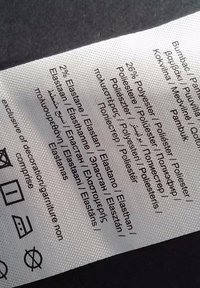 Etiqueta de cuidado de tejido blanco con texto negro que lista la composición del material: 2% elastano, 26% poliéster, múltiples traducciones. Incluye símbolos de cuidado.