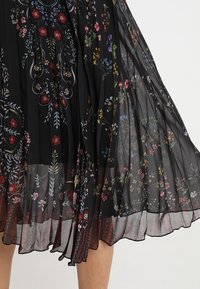 Jupe plissée noire avec un motif floral ornée de fleurs brodées colorées, fabriquée en tissu léger et transparent avec un design superposé.