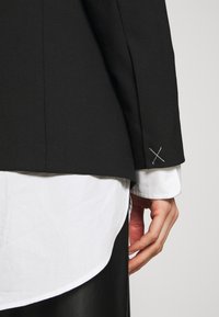 Černý propracovaný sako s hladkou texturou, s jedinečnými detaily na rukáve v podobě překřížených bílých stehů, přes bílé dress shirt.