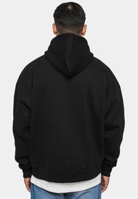 Sweatshirt à capuche noire avec une coupe décontractée, des poignets et un ourlet côtelés, fabriqué en tissu doux, porté par-dessus une chemise blanche superposée.