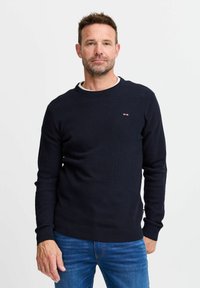 Marineblå strikkegenser med rund hals, teksturert stoff, ribbestrikkede mansjetter og nederkant. Lite logoakzent på brystet i rødt, hvitt og blått.