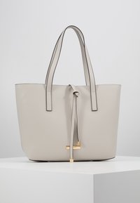 Beige læder tote taske med to lange håndtag, en knyttet lukning og en guldfarvet detalje i bunden, der har en glat overflade og minimalistisk design.