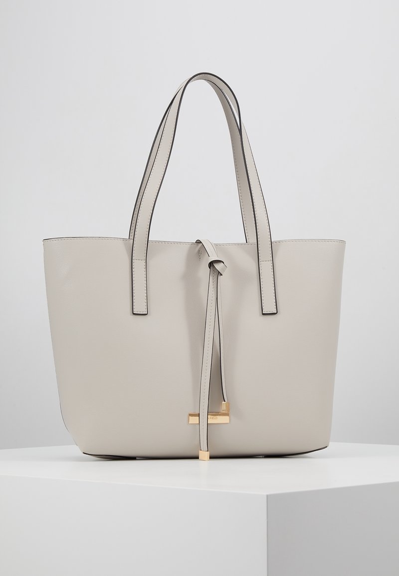 Beige læder tote taske med to lange håndtag, en knyttet lukning og en guldfarvet detalje i bunden, der har en glat overflade og minimalistisk design.